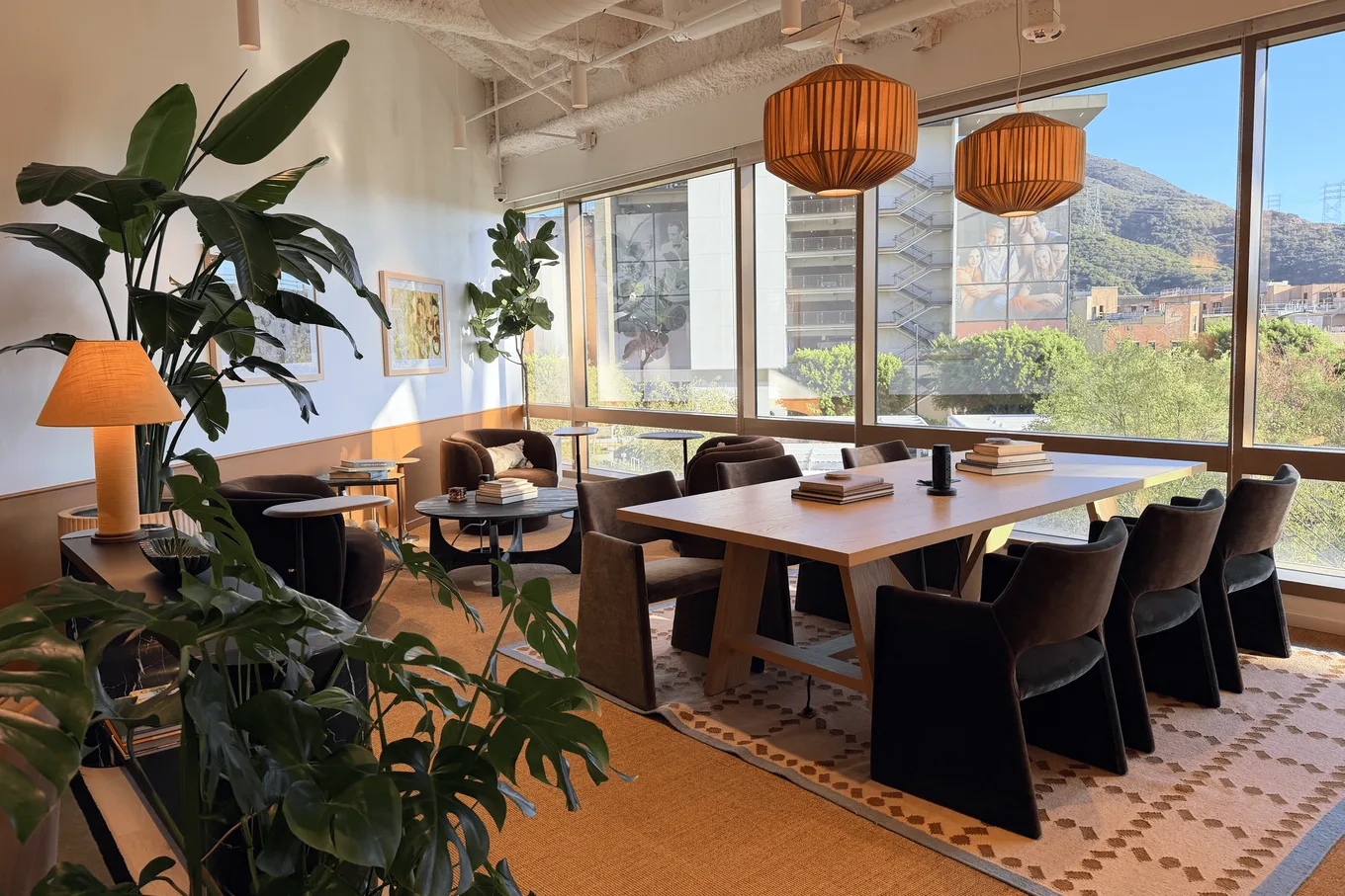 A sunlit studio meeting room: a long oak table under rattan pendants, velvet chairs, large plants and a wall of windows onto the hills
